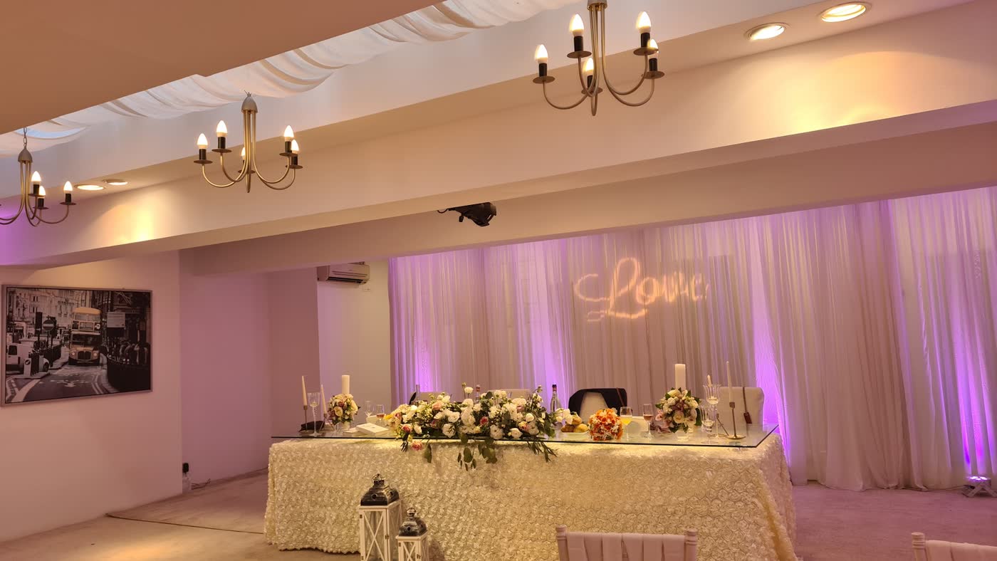 Sala interioară: draperii albe luminate în roz, candelabre și masa principală cu flori, sub cuvântul Love proiectat pe perete